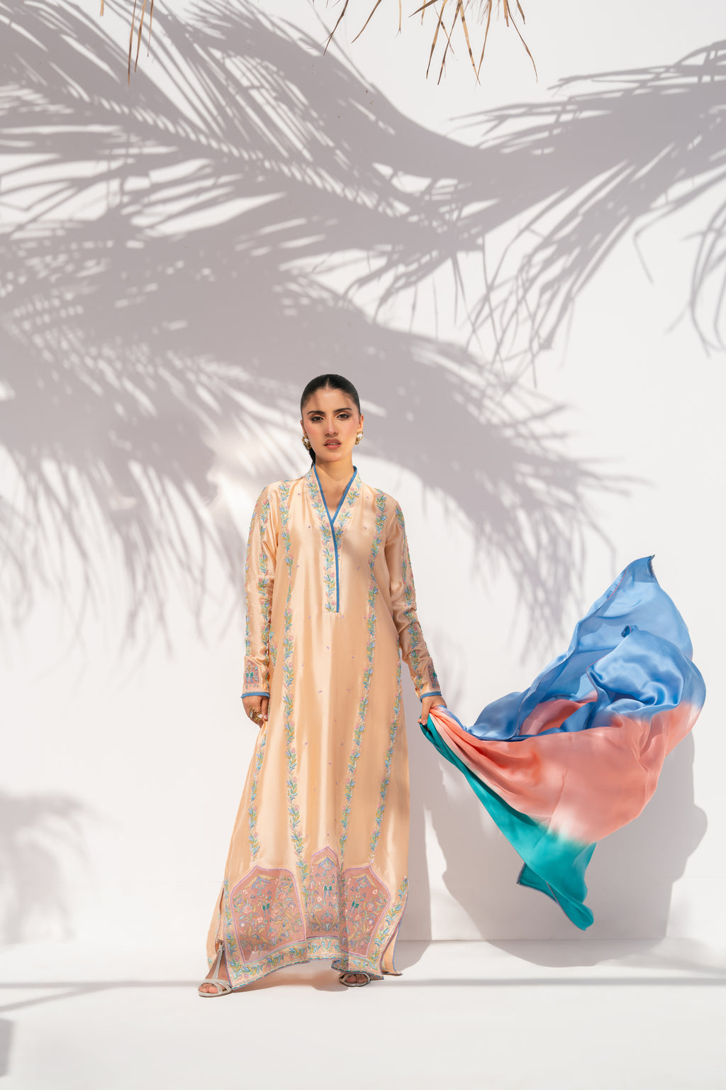Woman in a long, light-colored dress holding a colorful fabric against a white background with palm leaf shadows.