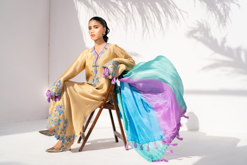 Woman in a traditional outfit with floral patterns and a colorful scarf, sitting on a chair.