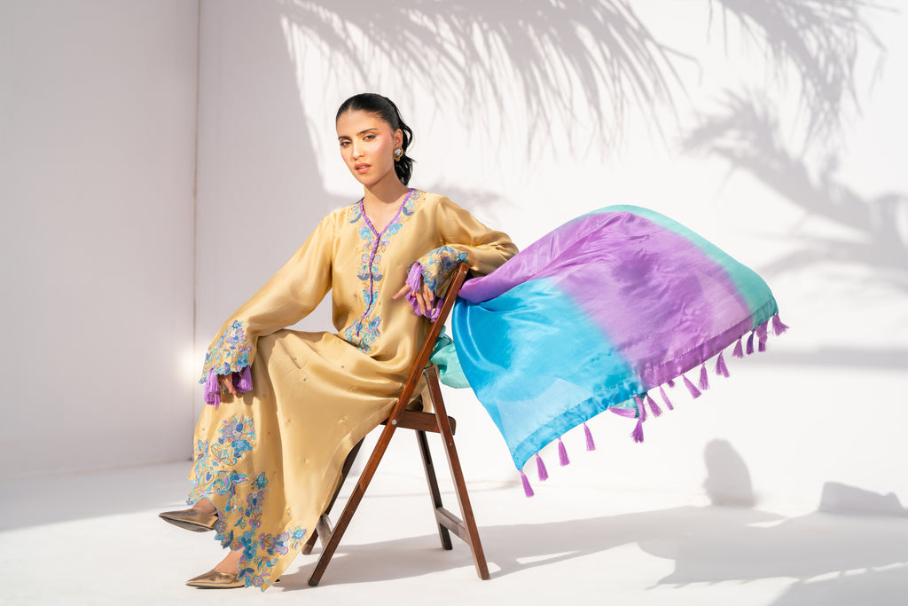 Woman in a yellow embroidered dress sitting on a chair with a colorful scarf, against a white background.